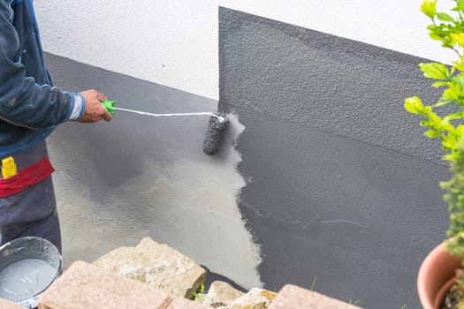 Close-up arm of a painter with paint roll in hand paints a house wall with gray paint.
