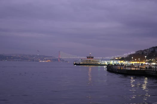 A ferry rests in the water at dusk with a bridge glowing in the distance.