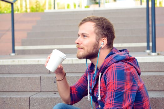 young bearded man in checkered shirt sitting on stairs listening music and drinking soft drink