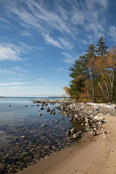 Katherine cove and the pines there inspired the group of seven artists. Image shows dead calm clear lake with rocks, sandy beach and fall trees. Blue sky with directional cirrus clouds