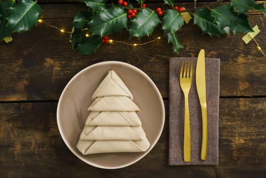 Christmas place setting with golden silverware, holly, fairy lights and a napkin folded as a Christmas tree