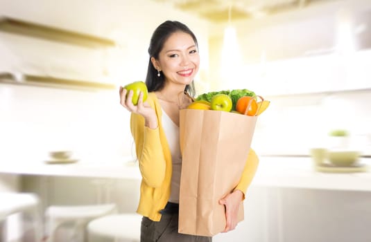 Young Asian female hand holding shopping paper bag filled with fruits and vegetables in kitchen.