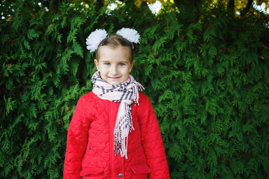 Smiling baby girl in a red jacket with white bows on her head. Child posing on a background of green arborvitae shrubs.