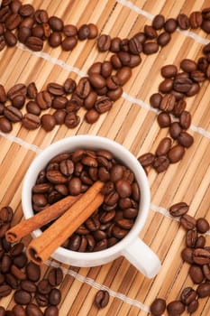 Coffee cup filled with coffee beans and two cinnamon sticks on bamboo mat with ornaments of coffee beans.