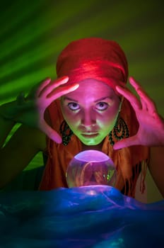 Older female teenager as a fortune teller in gypsy garb in front of a glass ball colorfully illuminated in a dark appearance.