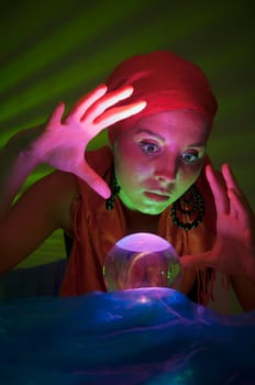 Older female teenager as a fortune teller in gypsy garb in front of a glass ball colorfully illuminated in a dark appearance.
