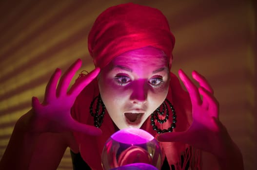 Older female teenager as a fortune teller in gypsy garb in front of a glass ball colorfully illuminated in a dark appearance.