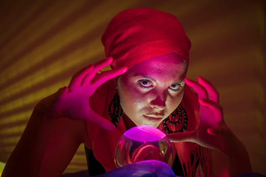 Older female teenager as a fortune teller in gypsy garb in front of a glass ball colorfully illuminated in a dark appearance.