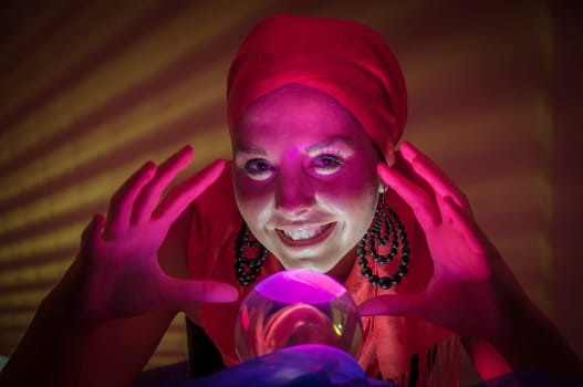 Older female teenager as a fortune teller in gypsy garb in front of a glass ball colorfully illuminated in a dark appearance.