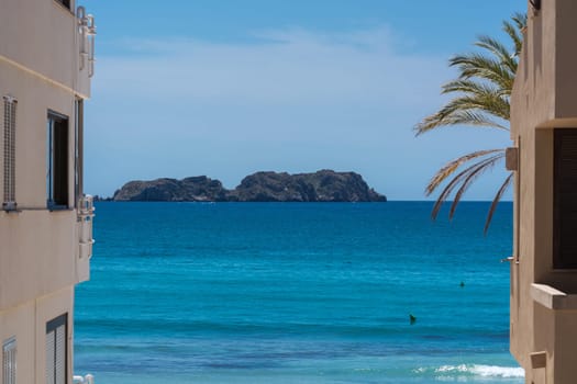 View of the blue sea in the foreground a House in Spanish style and a palm tree..