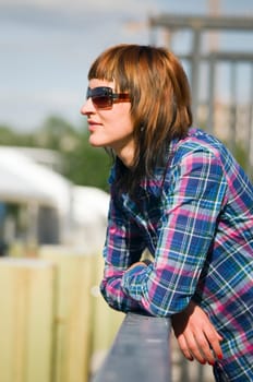 Side upper body view of a young brunette woman with plaid shirt and sunglasses leaning on a railing and looking to the side