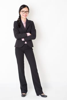 Full body portrait of young Asian businesswoman in formalwear arms folded and smiling, standing on plain background.