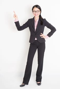 Full body portrait of young Asian businesswoman in formalwear fingers pointing at copy space and smiling, standing on plain background.