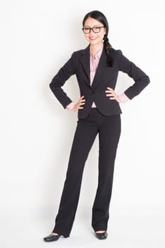 Full body portrait of young Asian businesswoman in formalwear hands on waist and smiling, standing on plain background.