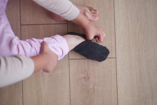 A child is pulling on a black sock while seated on a wooden floor.