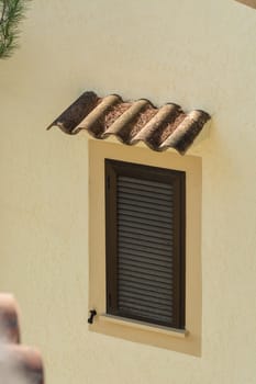 Window with brown closed shutter. Rain cover made of roof tiles in a yellow painted house wall..