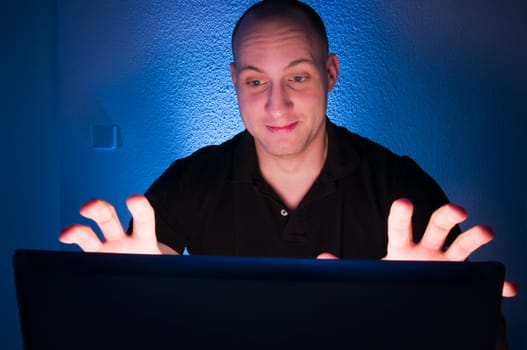 Head-and-shoulder shot of a young man sitting on a laptop monitor against a nightly blue background with a shocked expression on his face and frightened raised hands towards the monitor Camera position is behind the monitor towards the man's face