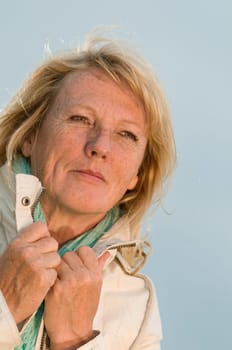 Upper body portrait of a blonde mature woman in a light jacket against an overcast sky looking dreamily to the side with her hands on the collar  Increased view