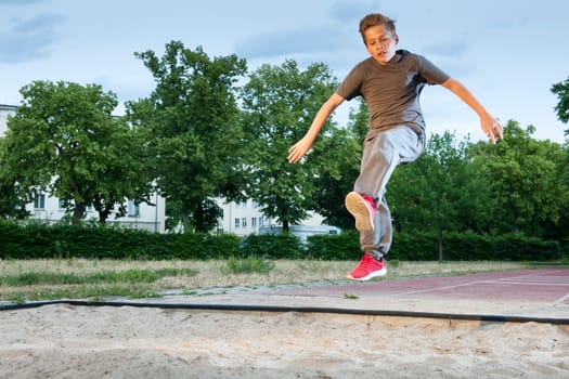 Side frontal full-body view of a male young teenager frozen while jumping into a long jump pit.