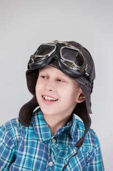 Against a grey background,the exposed upper body view of a ten-year-old boy in a blue-checkered short-sleeved shirt and brown leather cap with classic motorcycle glasses looks happily to the side.