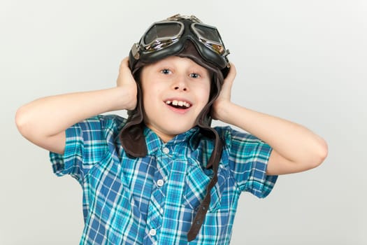 Against a grey background,the exposed upper body view of a ten-year-old boy in a blue-checkered short-sleeved shirt and brown leather cap with classic motorcycle glasses smiling joyfully into the camera.