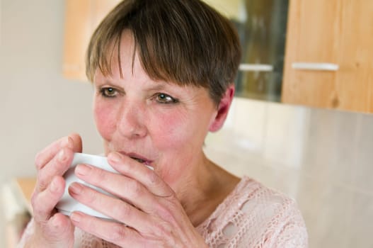 Side head-and-shoulder portrait of an elderly woman drinking from a white cup in a home environment.