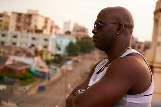 Black gang member with arms crossed contemplating cityscape at sunset.