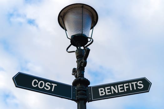 Street lighting pole with two opposite directional arrows over blue cloudy background. Cost versus Benefits concept.