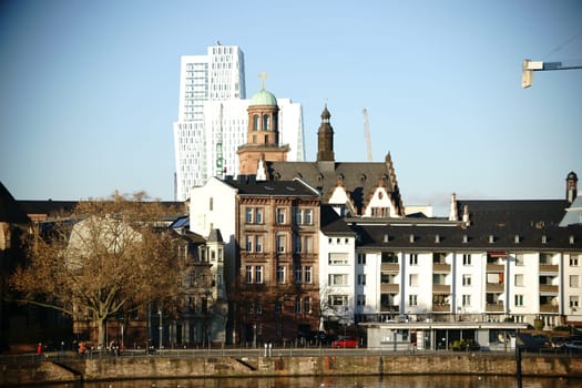 The tower of the Church of Our Lady and other building towers behind residential buildings on the banks of the Main in Frankfurt.