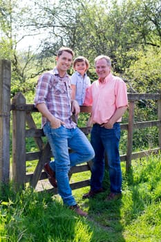 A little boy is sitting on a farm gate in the countryside with his father and his grandfather. They are all smiling at the camera.