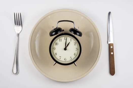 On the plate is an alarm clock, lying next to a knife and fork, white background, top view