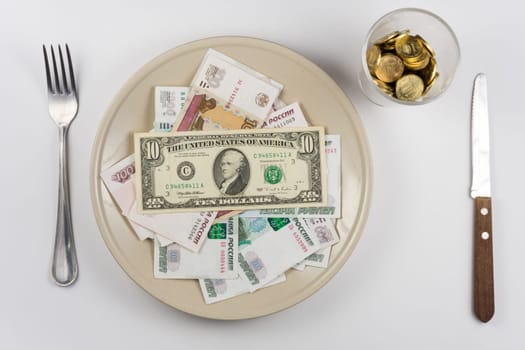 On the plate is a lot of money Russian ruble and the US dollar, standing next to a glass and a fork and knife lie, top view