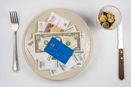 On the plate is a lot of money and a plastic credit card, standing next to a glass and a fork and knife lie, top view