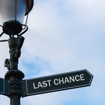 Street lighting pole with conceptual message LAST CHANCE on directional arrow over blue cloudy background.