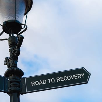 Street lighting pole with conceptual message ROAD TO RECOVERY on directional arrow over blue cloudy background.