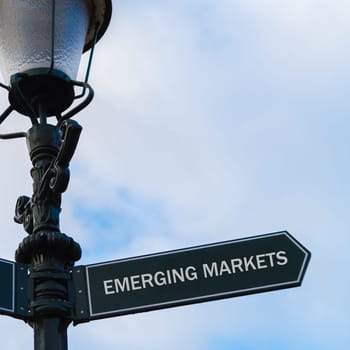 Street lighting pole with conceptual message EMERGING MARKETS on directional arrow over blue cloudy background.