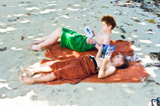 brothers are lying at the sandy beach in the shadow, relaxing and reading books