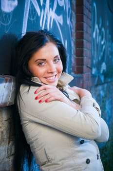 Upper body view of a young black-haired woman in a trench coat with a high collar leaning against a graffiti sprayed brick wall.