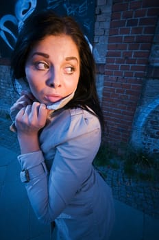 Young black-haired woman on a brick wall sprayed with graffiti in the evening illuminated with artificial light looking back anxiously.