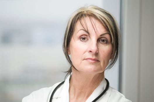 Frontal head-and-shoulder portrait of a middle-aged woman in a white coat with a stethoscope standing in front of a window and looking seriously into the camera.
