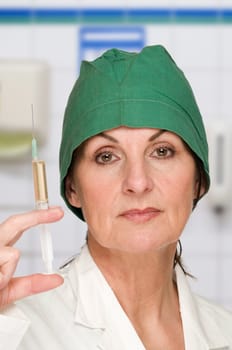 Head-and-shoulder view of a middle-aged woman with a green surgical hood against a white background holds a filled syringe in front of her.
