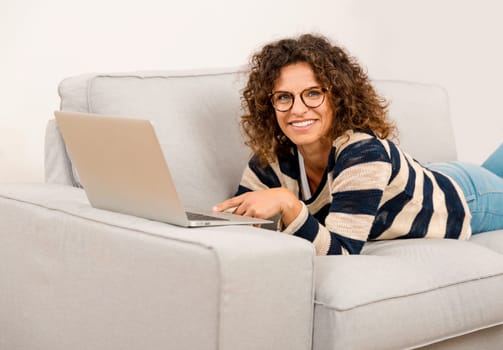 Beautiful woman at home lying on the sofa and working with a laptop