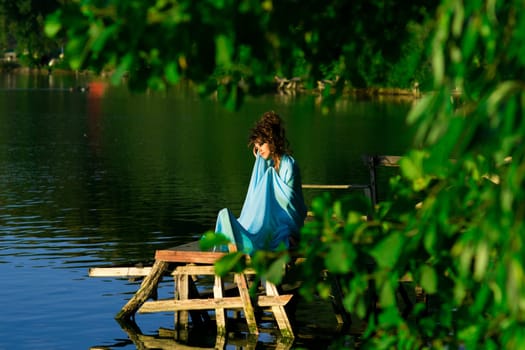 Extremely beautiful girl with perfect professional hairstyle.Nimf near water lying on the sun.Nymph,diva,goddess sits near the water,lake,river with blue dress,green trees,professional hairstyle. Summer greenery.Nimf near the water lying on the sun.