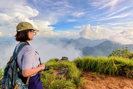 Hiker asian teens girl with camera and backpack wear caps and glasses looking beautiful landscape nature of mountain range and blue sky during sunset at Phu Chi Fa Forest Park, Chiang Rai, Thailand