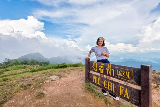 Tourist teen girl hiker wear cap and glasses standing smile pose thumb up near nameplate attractions of Phu Chi Fa Forest Park on mountain and blue sky background, Chiang Rai, Thailand