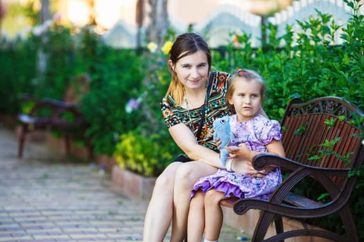 Mother and little daughter sit on a park bench on a background of bright green foliage. Hugging mother and daughter. Family portrait. Selective focus.