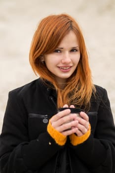 Beautiful red-haired girl in a black fashionable coat trying to keep warm in the cold weather.Cheerful smiling red-haired girl with cute smile with a cup of hot tea, coffee, chocolate.Cheerful smiling red-haired girl with cute smile red hot coffee.