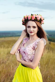 Beautiful fashionable attractive girl with big wreath of beautiful bright flowers on her hair, head. Sitting in field cute girl with big bouquet ful of many yellow flowers in summer. Blue sky, yellow field, many flowers.