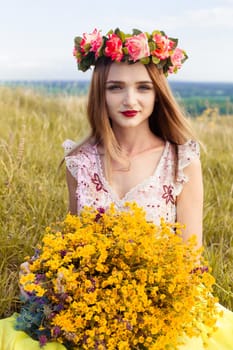 Beautiful fashionable attractive girl with big wreath of beautiful bright flowers on her hair, head. Sitting in field cute girl with big bouquet ful of many yellow flowers in summer. Blue sky, yellow field, many flowers.