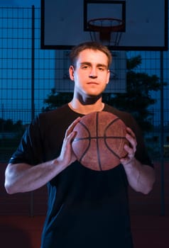 Frontal upper body view of a basketball player in the evening in the light with basketball held in front of him.